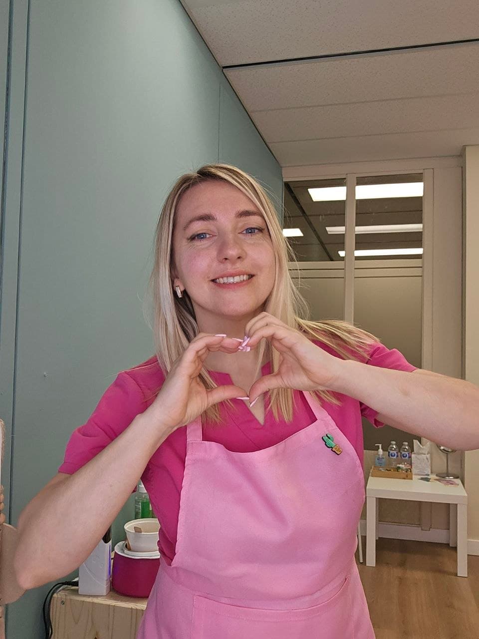 Elena, owner of Oh, Honey, in pink scrubs making a heart shape with her hands